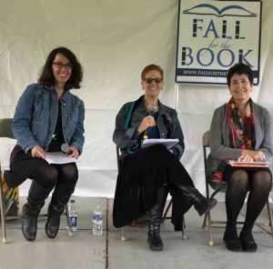 Three women sit on a stage. They are holding copies of a book they are reading from and the women in the middle holds a microphone.