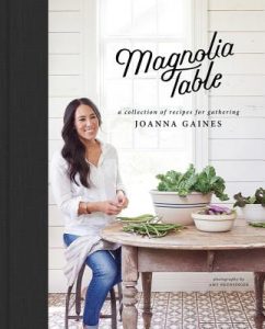 The cover of Magnolia Table shows Joanna Gains sitting at a kitchen table with a large bowl of greens beside her.