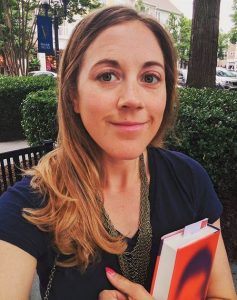 A woman with long blond hair holds a book with a red cover and smiles at the camera.