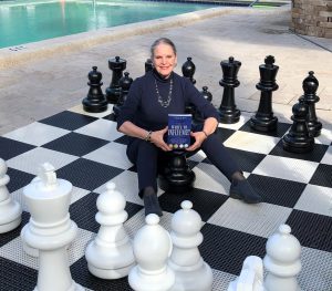 Carla Bass stands in the center of a giant chess set and holds her book, Write to Influence!