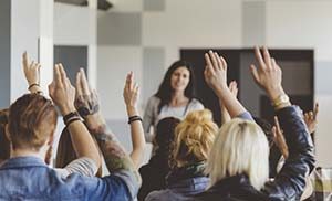 A woman speaking to a group. Almost everyone is raising their hand to ask a question.