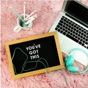 A silver laptop sits open next to sea green headphones, a mug full of chalk pens, and a small chalkboard that says "You've Got This."